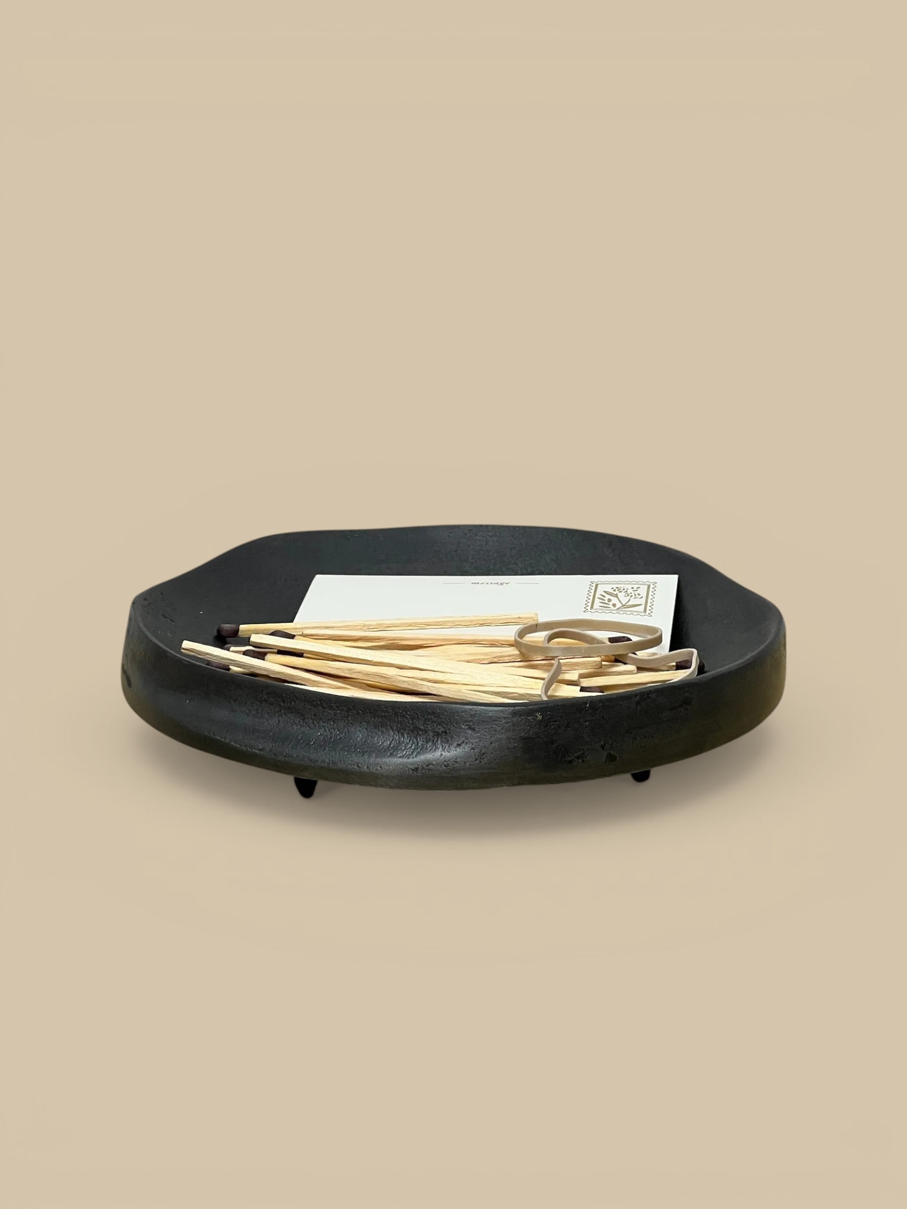Homestyle Collective matte black aluminum footed dish with organic shape, holding matches and a rubber band.