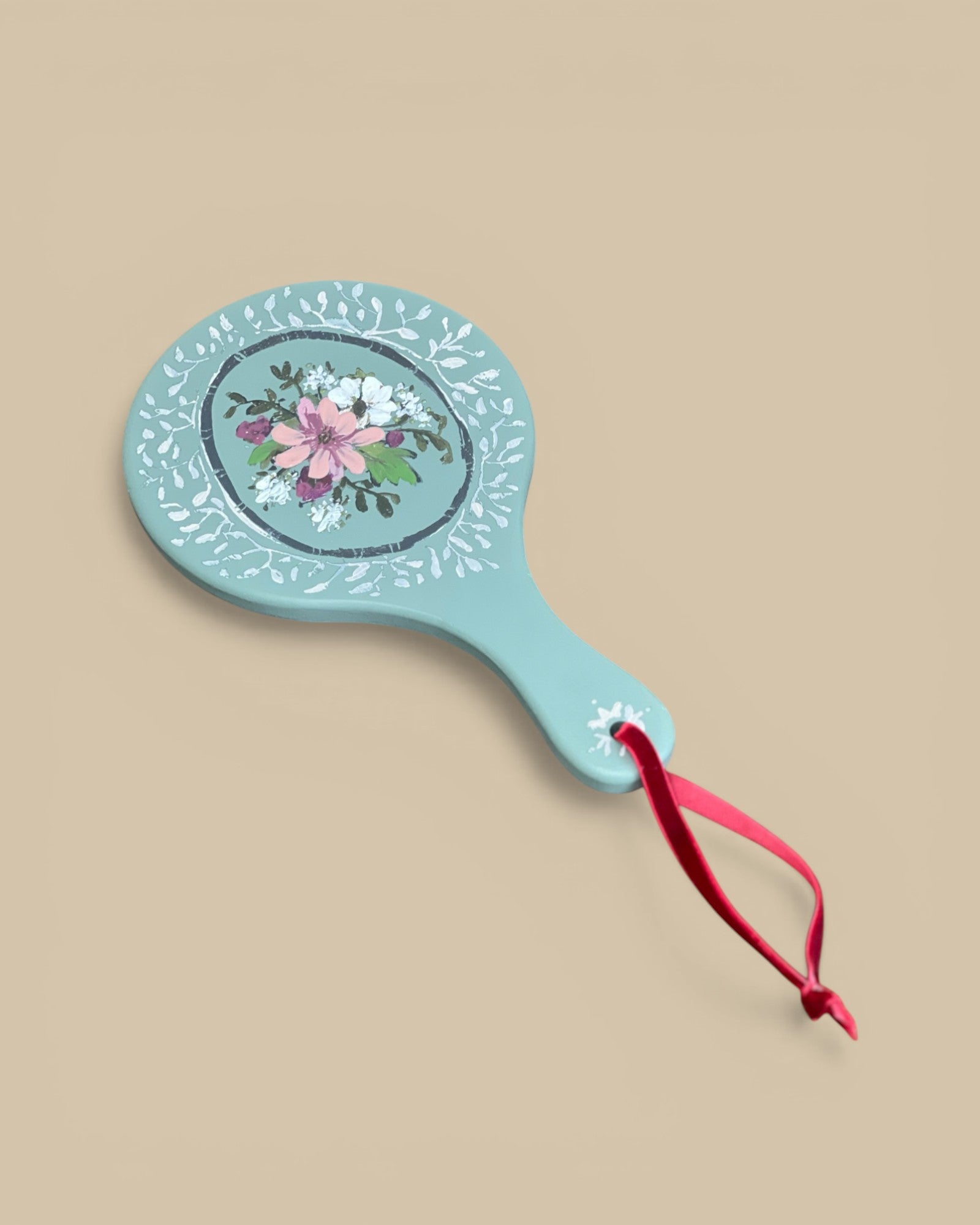 Homestyle Collective painted wood hand mirror with floral motif and red ribbon detail. Light blue with pink and white flowers.