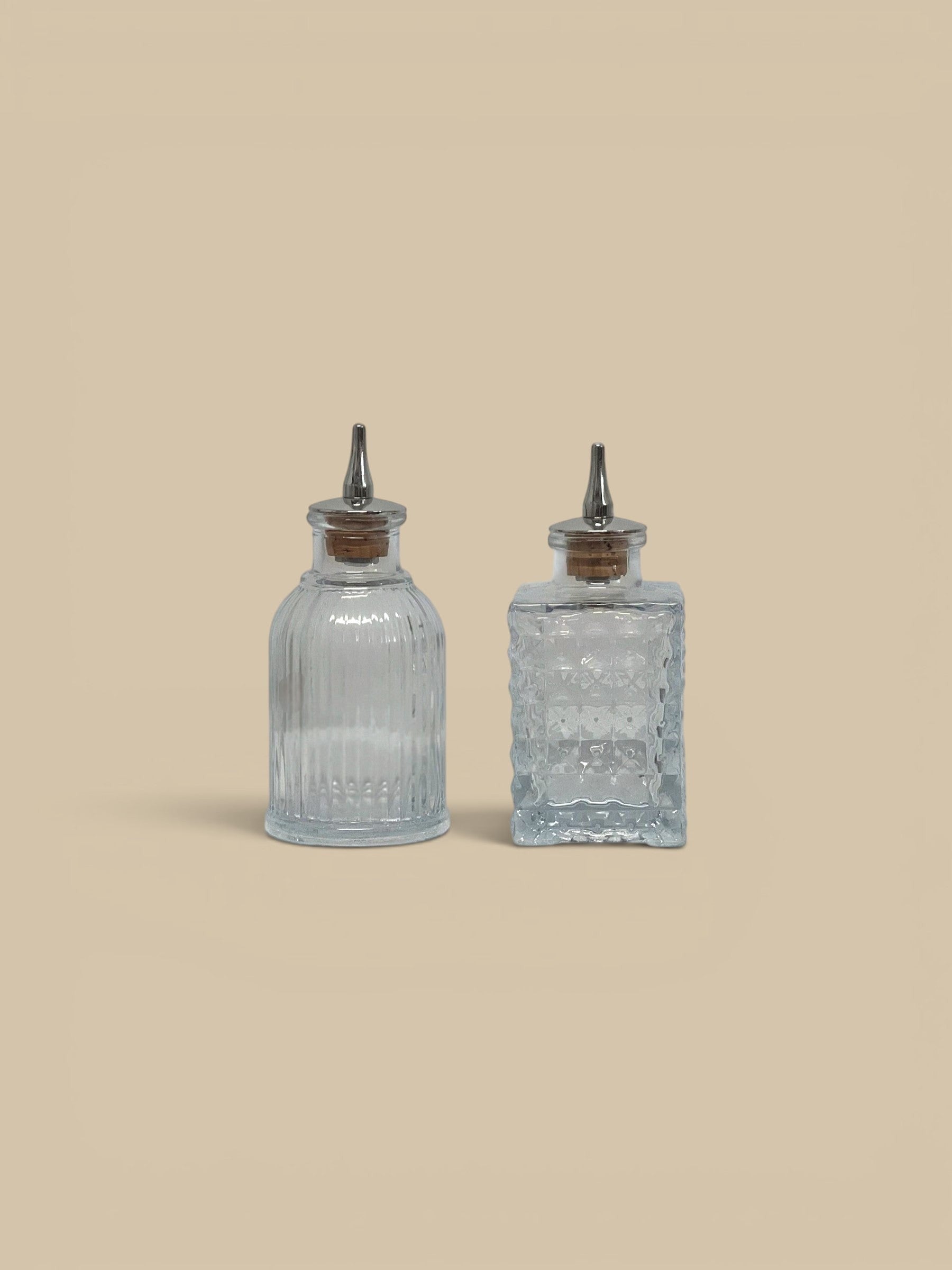 Homestyle Collective clear glass bitters bottles, one round with vertical ridges, one square with textured pattern, both with cork stoppers and pour spouts.