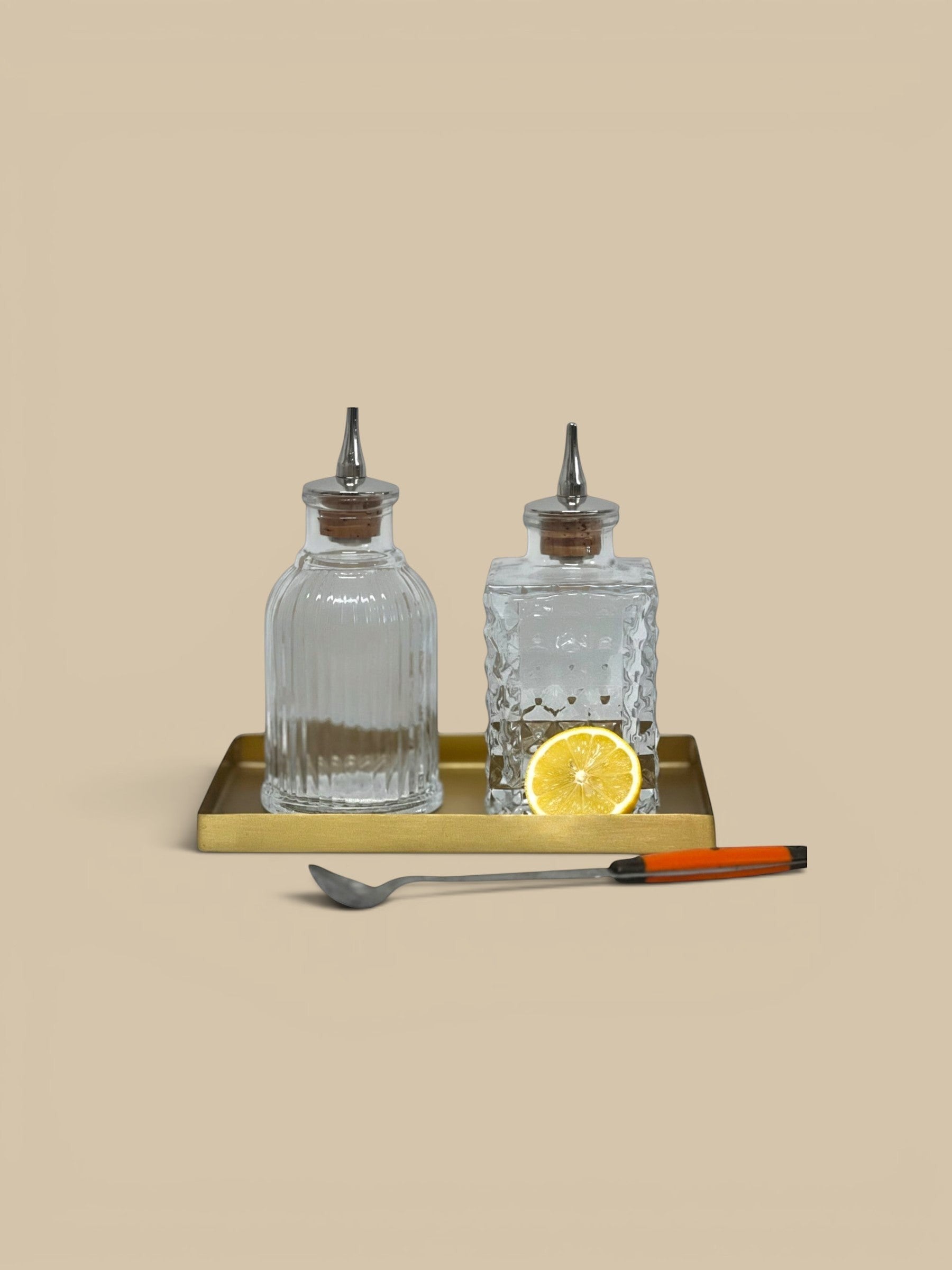 Homestyle Collective clear glass bitters bottles, one round ribbed and one square textured, with pour spouts on a gold tray with a spoon.