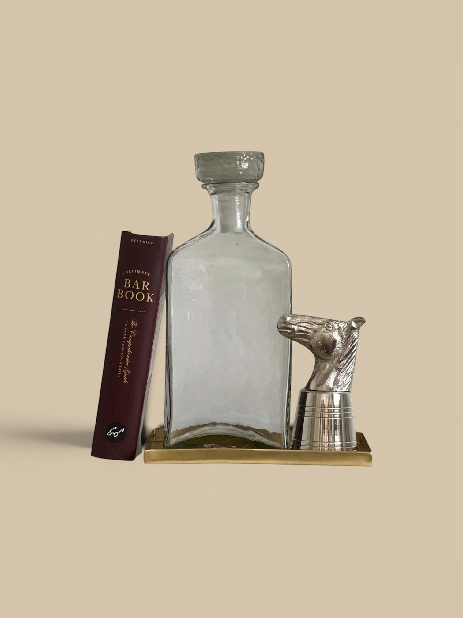 Homestyle Collective clear glass decanter with a textured grey stopper and a silver horse head stopper on a gold tray.