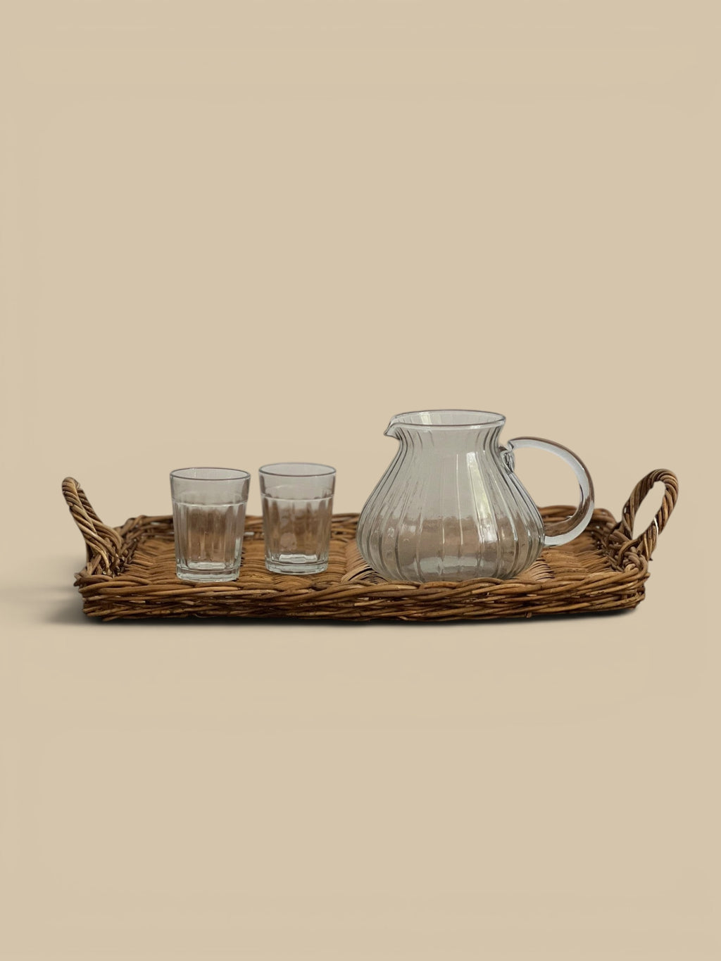 Homestyle Collective clear glass pitcher with ribbed texture and handle, beside two small glasses on a woven tray.