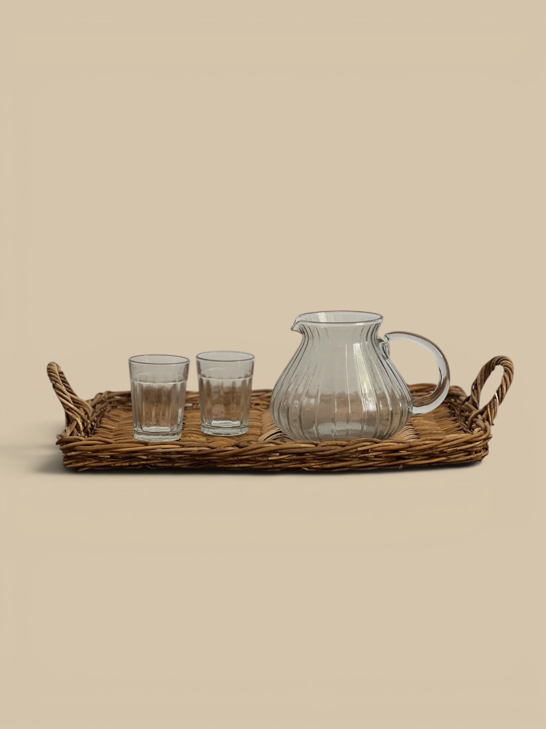 Homestyle Collective clear glass pitcher with ribbed texture and handle, beside two small glasses on a woven tray.