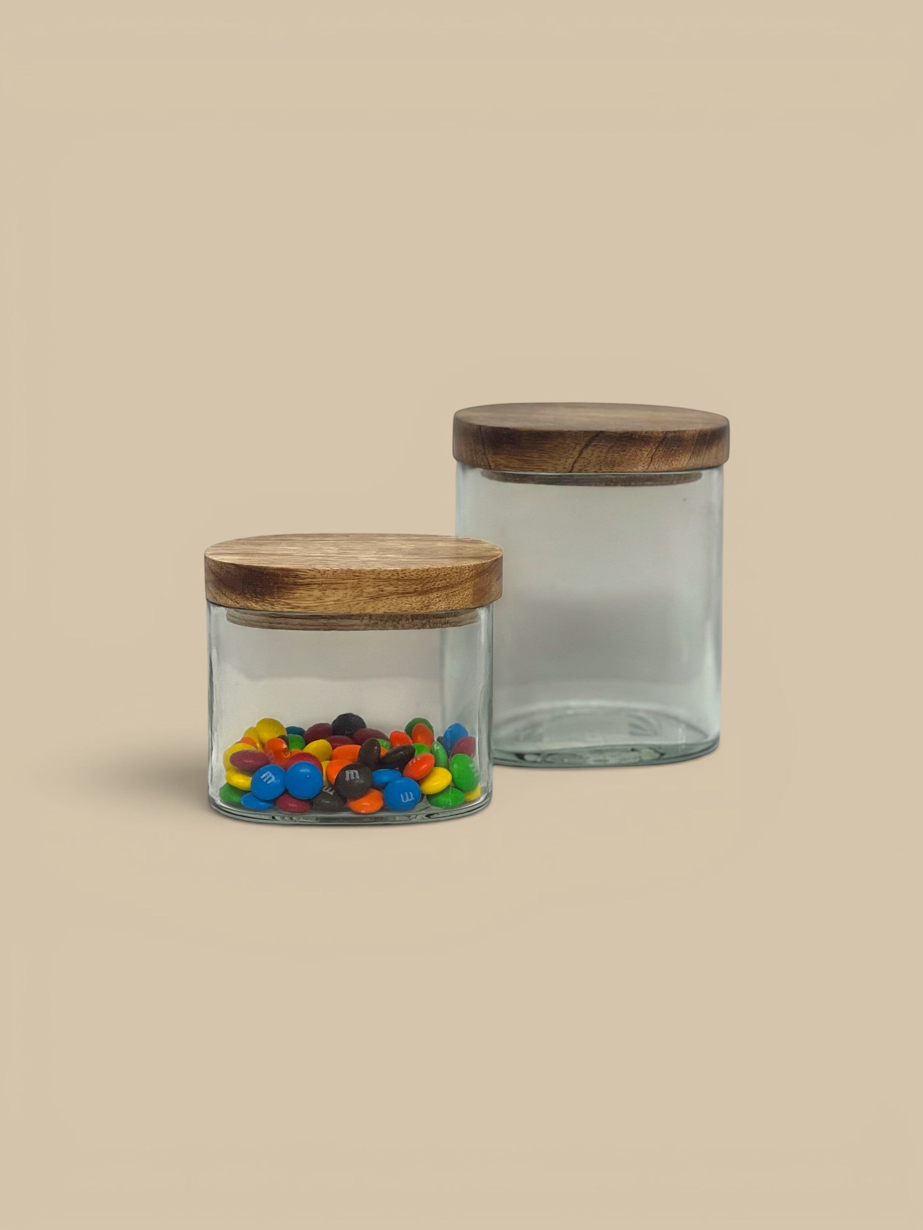 Homestyle Collective clear glass storage jars with natural wood lids, one filled with colorful candies.