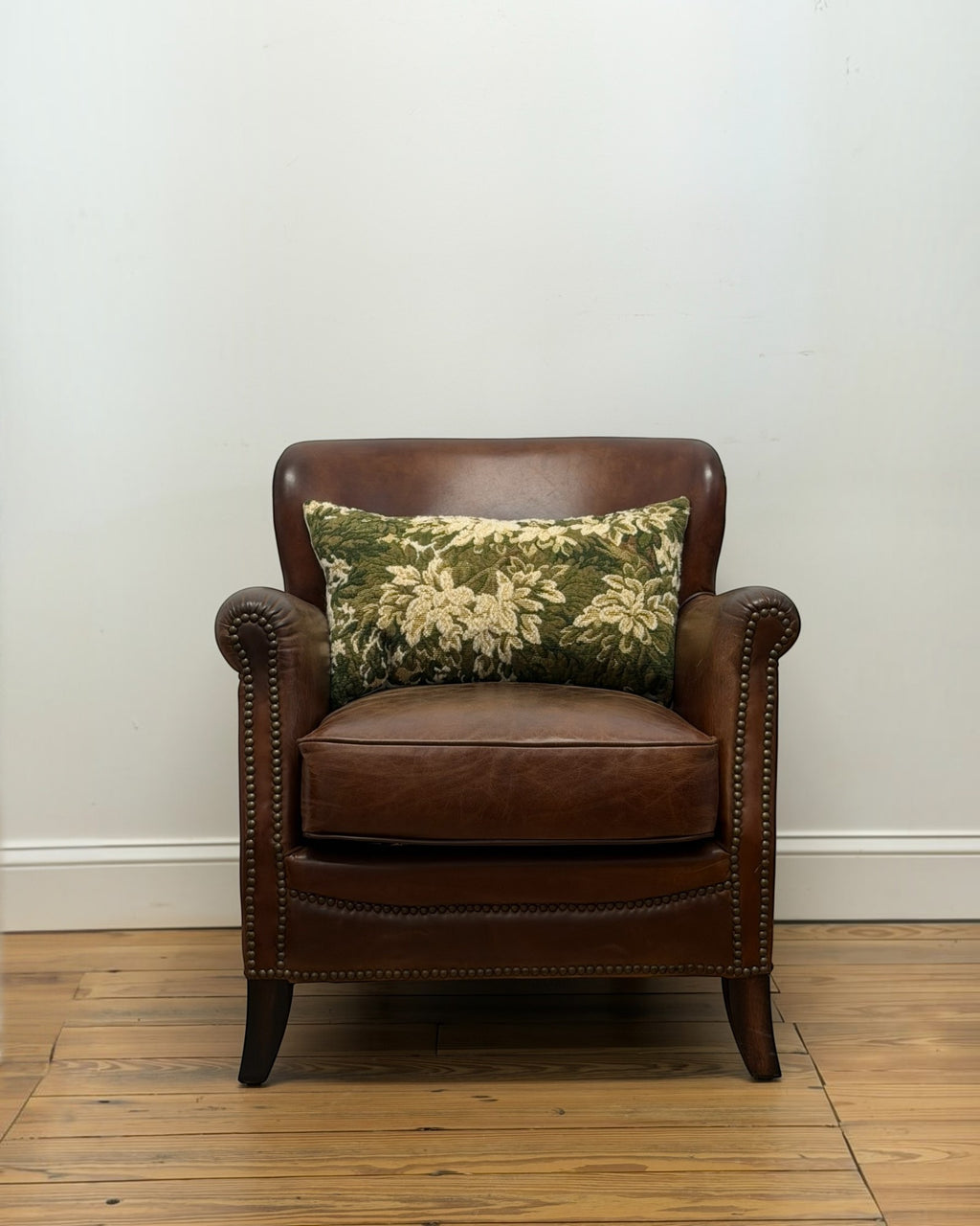 Homestyle Collective green floral tapestry throw pillow with vintage linen backing resting on a brown leather chair.