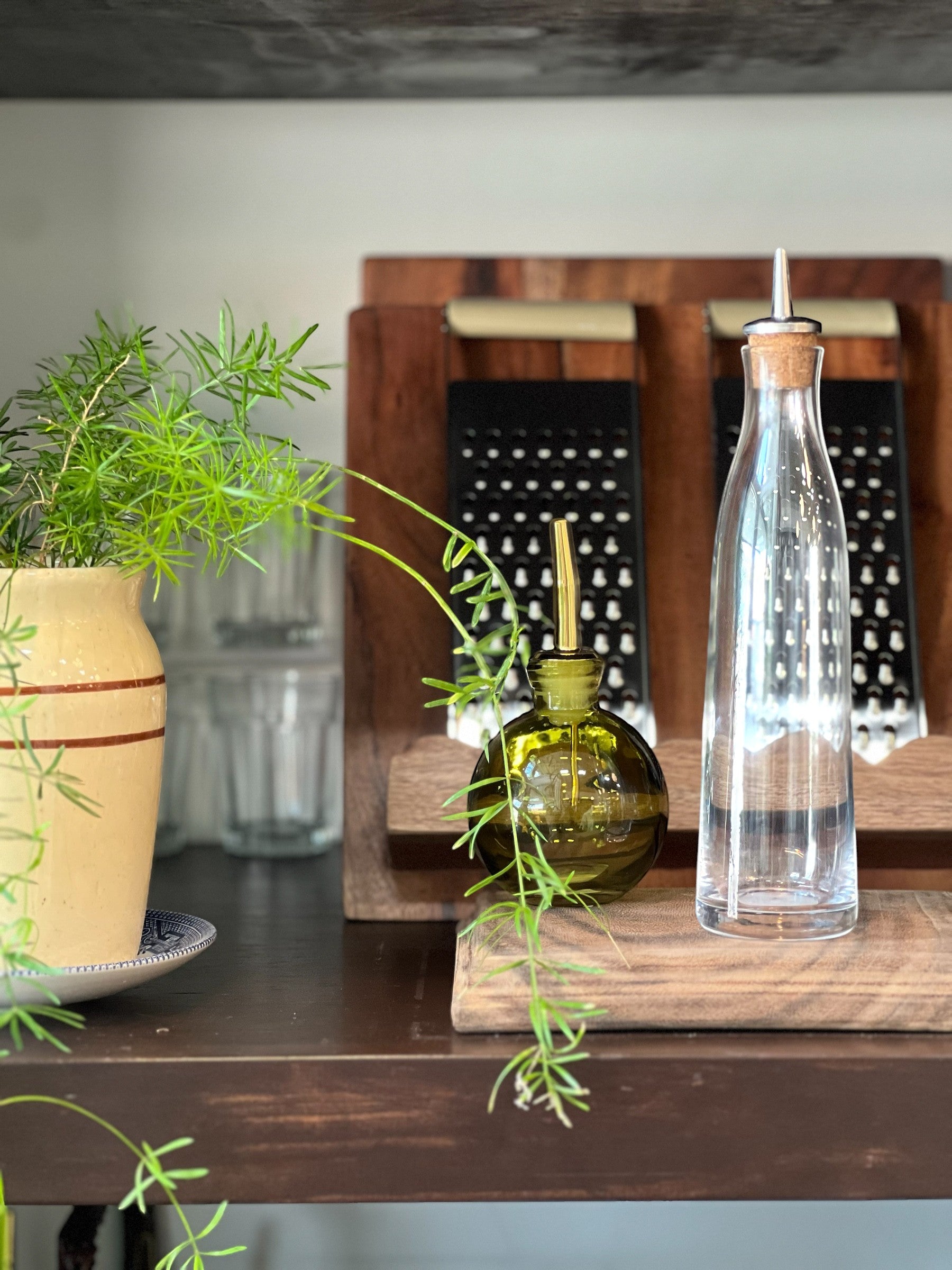 Homestyle Collective green glass oil cruet with gold spout and clear glass oil bottle with cork and metal spout on wooden board.