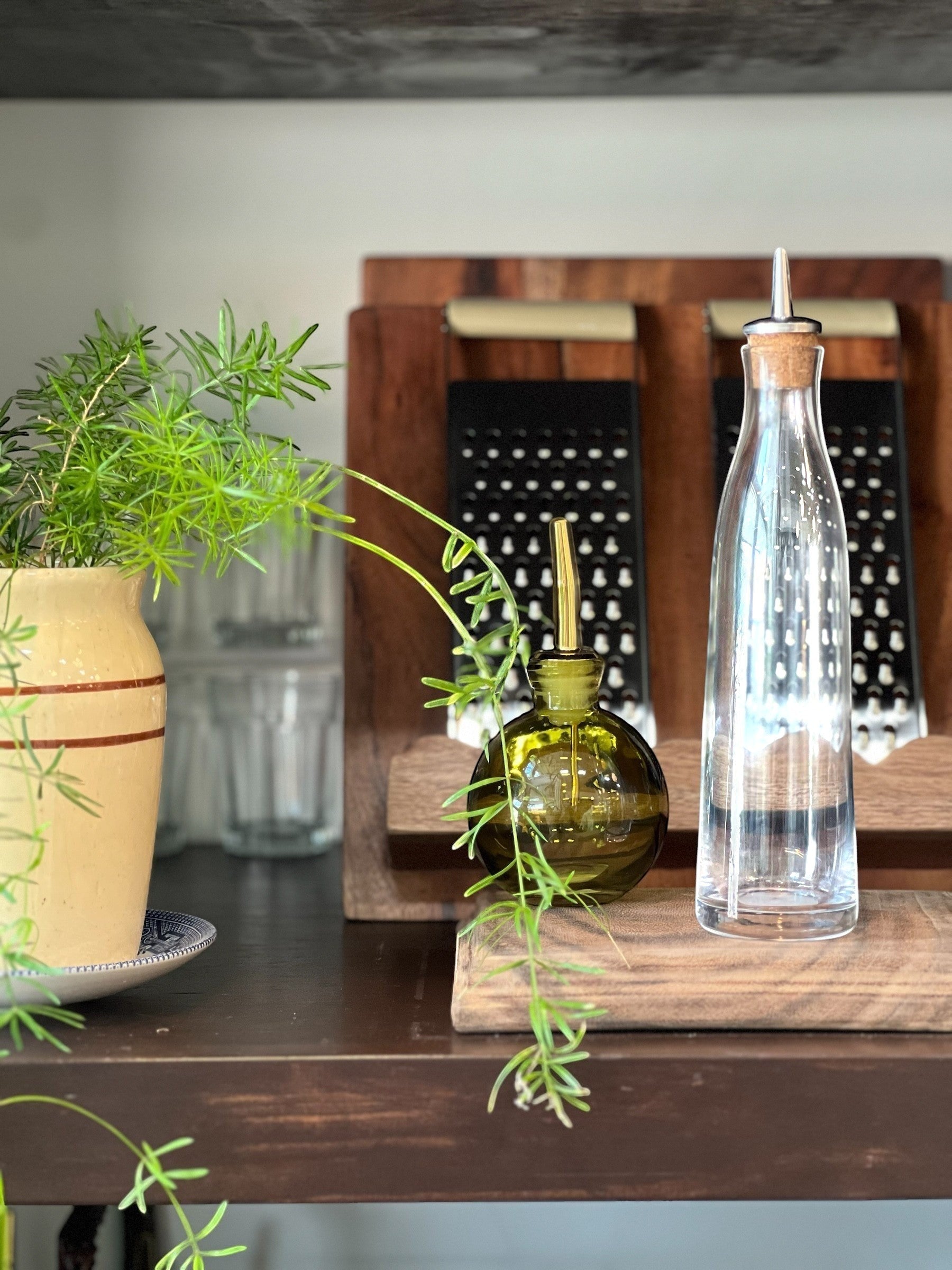 Homestyle Collective green glass oil cruet with gold spout and clear glass oil bottle with cork and metal spout on wooden board.