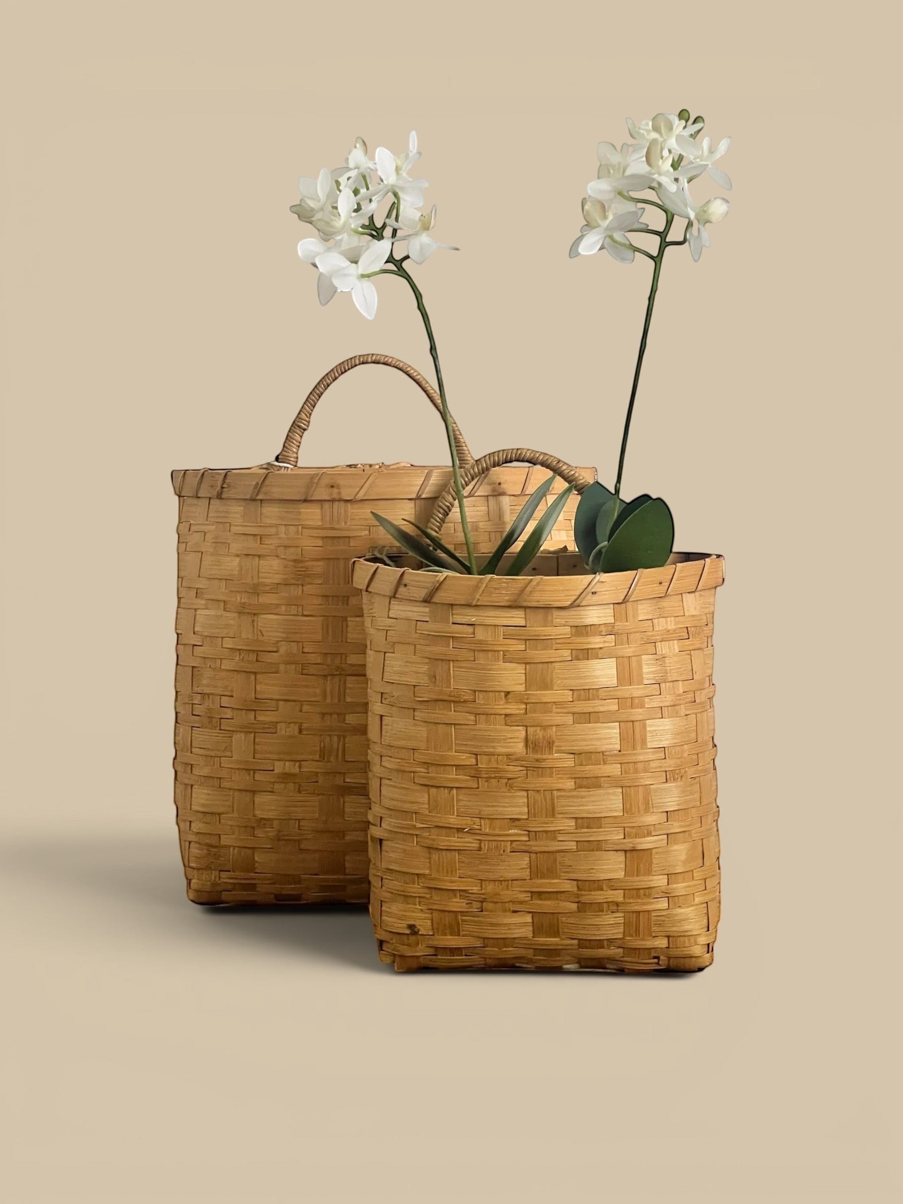 Homestyle Collective set of 2 natural rattan hanging baskets with built-in handles, one tall and flat, the other cylindrical, holding faux orchids.