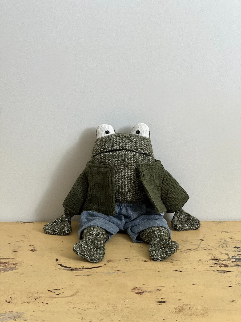 Homestyle Collective stuffed cotton frog toy wearing a green knit cardigan and blue shorts, sitting on a wooden surface.