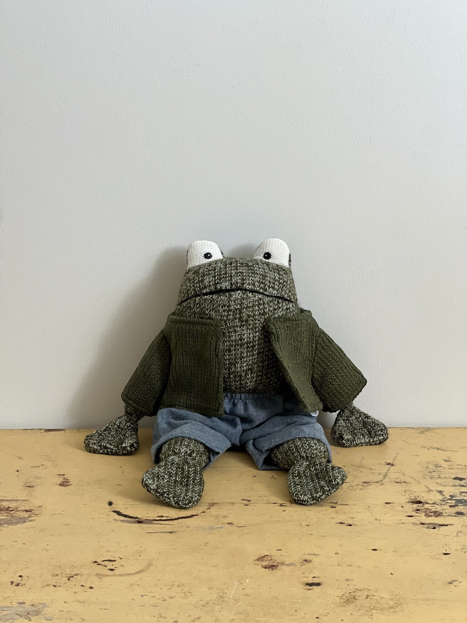 Homestyle Collective stuffed cotton frog toy wearing a green knit cardigan and blue shorts, sitting on a wooden surface.