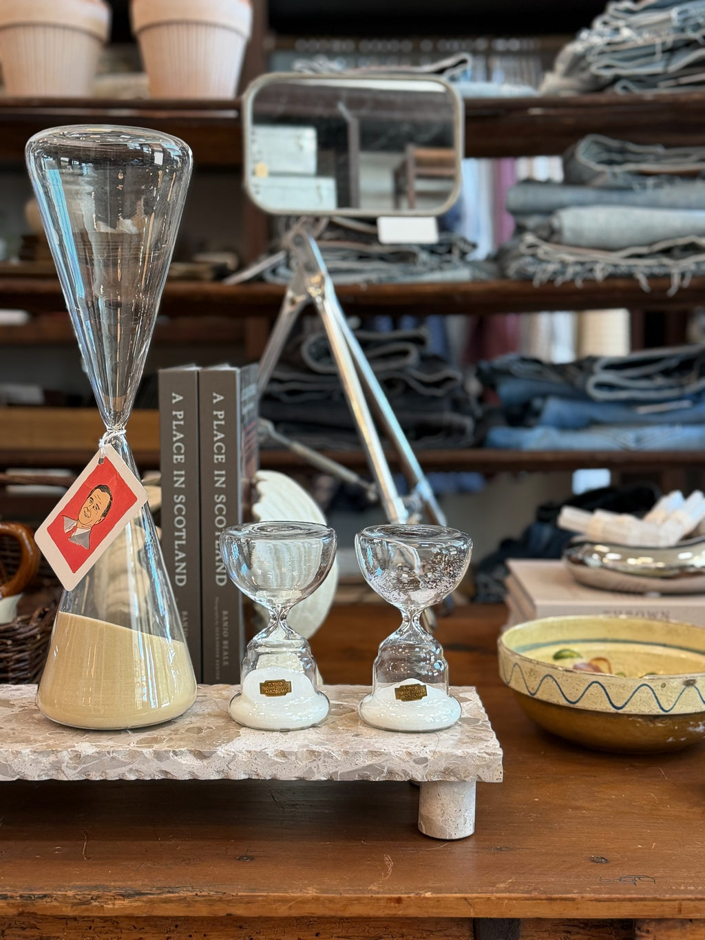Homestyle Collective trophy-shaped glass hourglass with tan sand on a stone display stand next to two smaller timers.