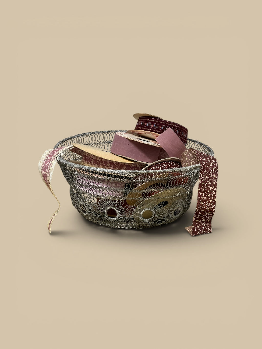 Homestyle Collective silver twisted wire basket with decorative circular cutouts, filled with spools of ribbon in burgundy, pink, and cream.