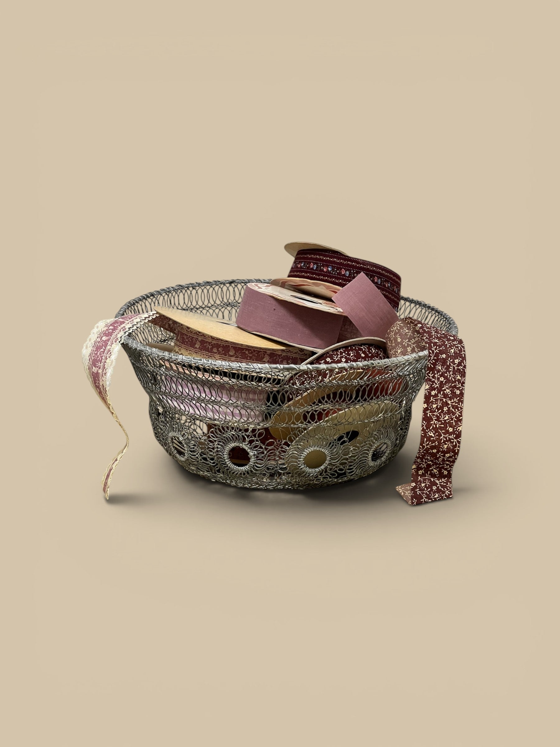 Homestyle Collective silver twisted wire basket with decorative circular cutouts, filled with spools of ribbon in burgundy, pink, and cream.