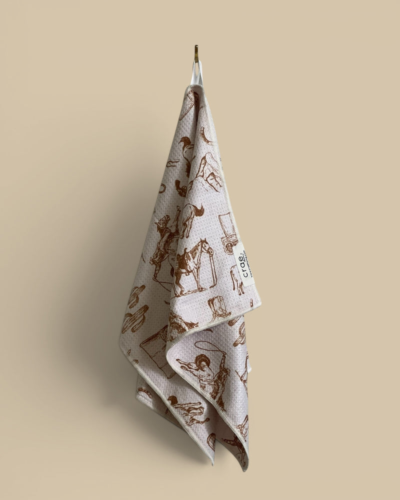 Homestyle Collective light pink waffle weave hand towel with brown cowboy and western-themed print.