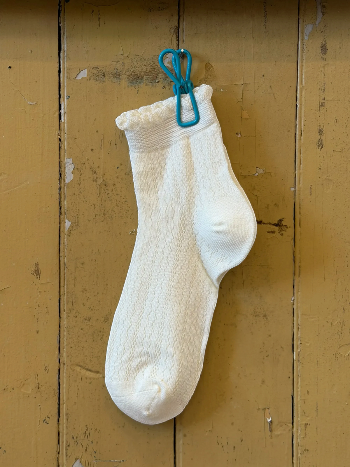 Homestyle Collective white mesh ankle socks with a delicate ruffle cuff, hanging from a blue clip on a wooden wall.