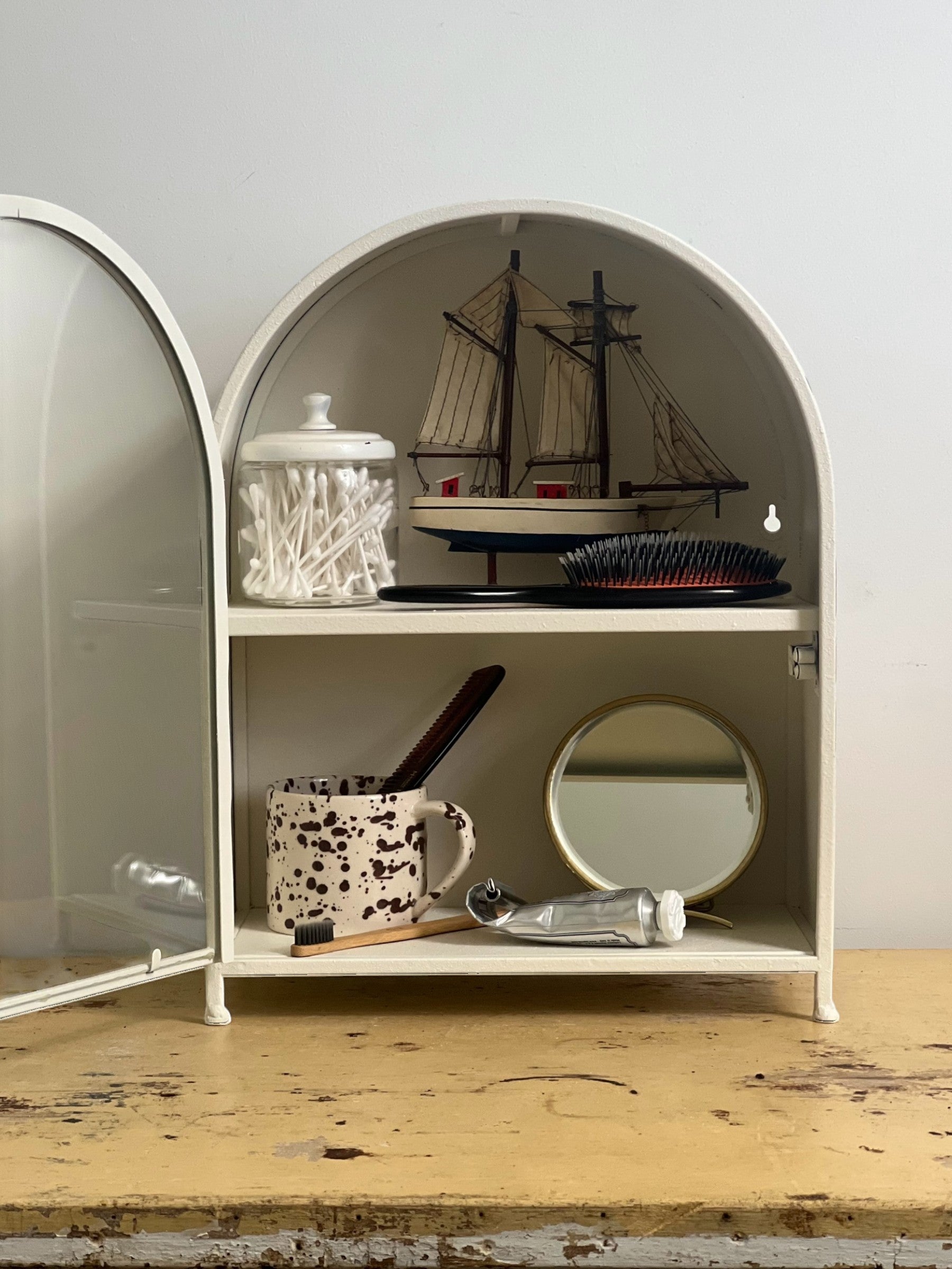 Homestyle Collective textured white metal arched display cabinet with glass door, featuring q-tips, model ship, brush, mug, mirror, and toothpaste.